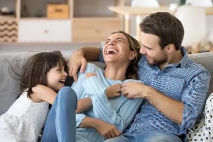 Happy family laughing together on a sofa at home: dad tickles mom while their young daughter giggles during a playful moment, reflecting the warmth and care provided by your trusted Family and Cosmetic Dentist in Dublin.